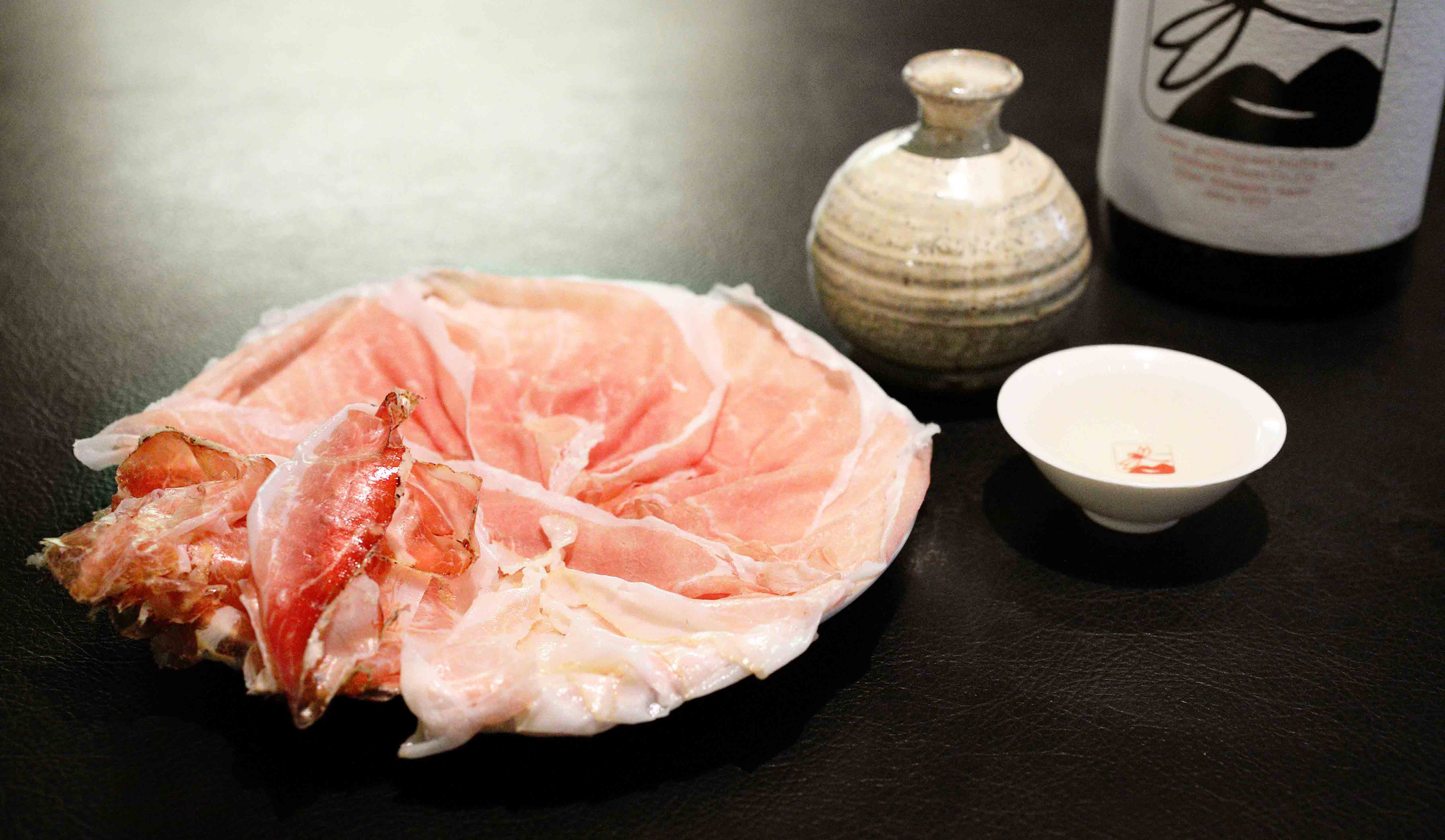 深夜のカウンター飯にちょうどいい生ハムの3種盛り合わせ。1800円。今宵は、柔らかく口どけのいい“プロシュット・ディ・パルマ 24ヶ月熟成”、肩ロースの生ハム、”ローザ・ディ・ノルチア”、スパイシーな香りと脂の旨みの調和が見事な、豚に頬の生ハム、”グアンチャーレ”。合わせた燗酒は、いづみ橋 生もと純米 黒トンボ。一合とっくり¥950。