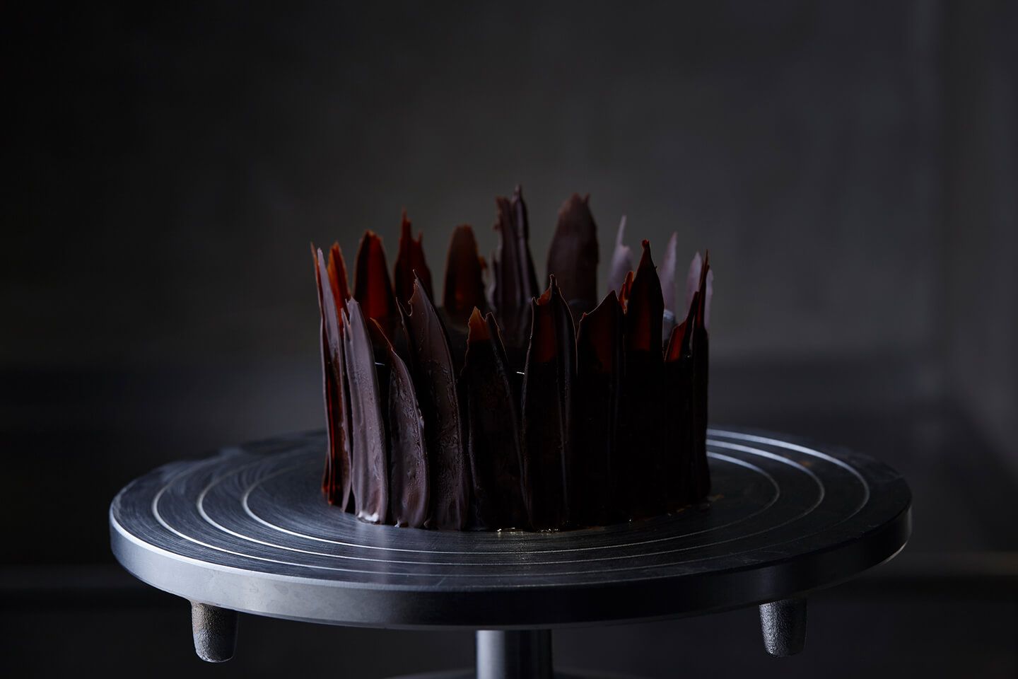 銀色のケーキ代の上に、ケーキの周囲にチョコレートを縦に塗り、王冠のように飾り付けたクーロンヌ ノアールがある