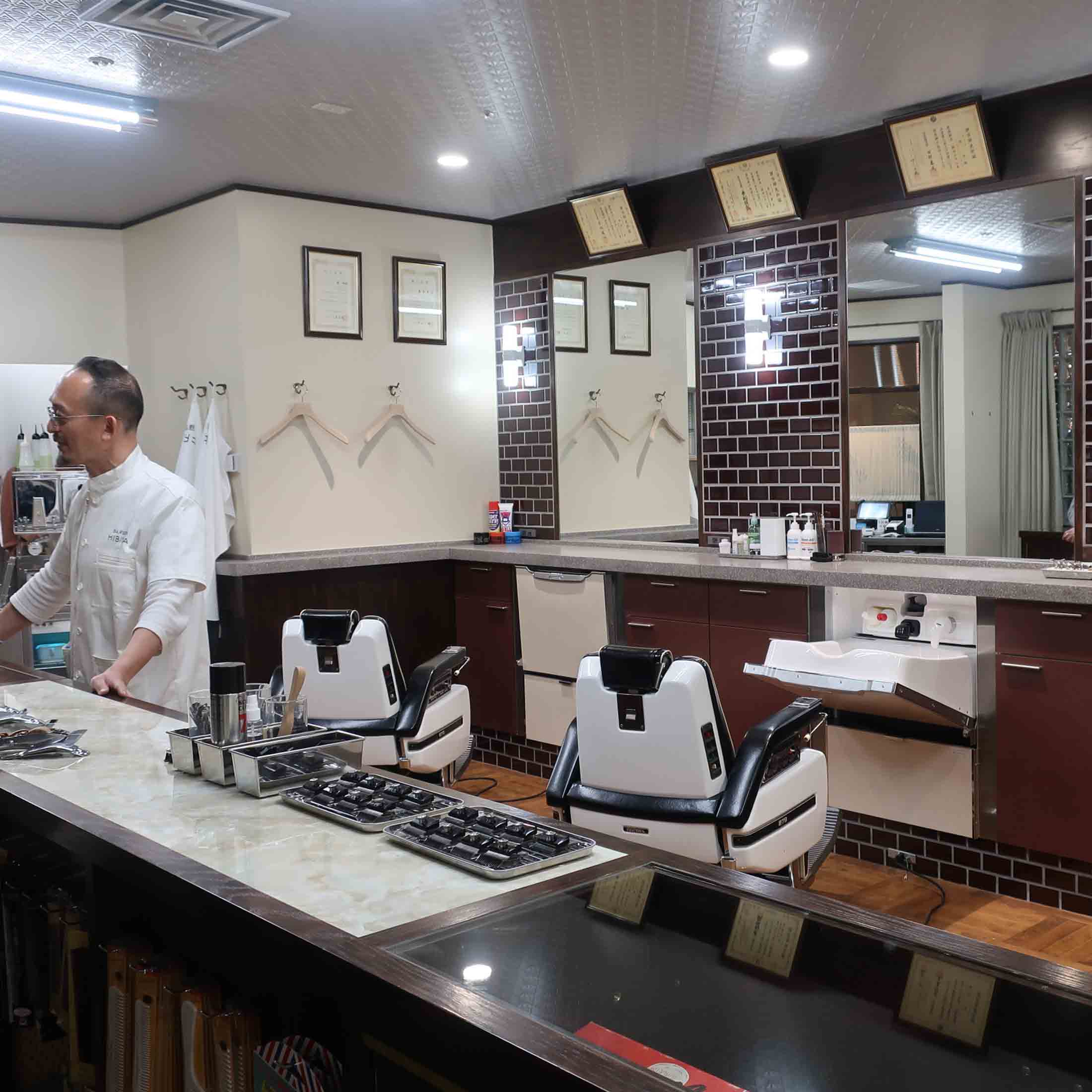 「理容 ヒビヤ」の店内。古き良き理容店の雰囲気。白いユニフォームの男性は店主の藤井実さん。蛍光灯調の照明もまた昭和な印象。