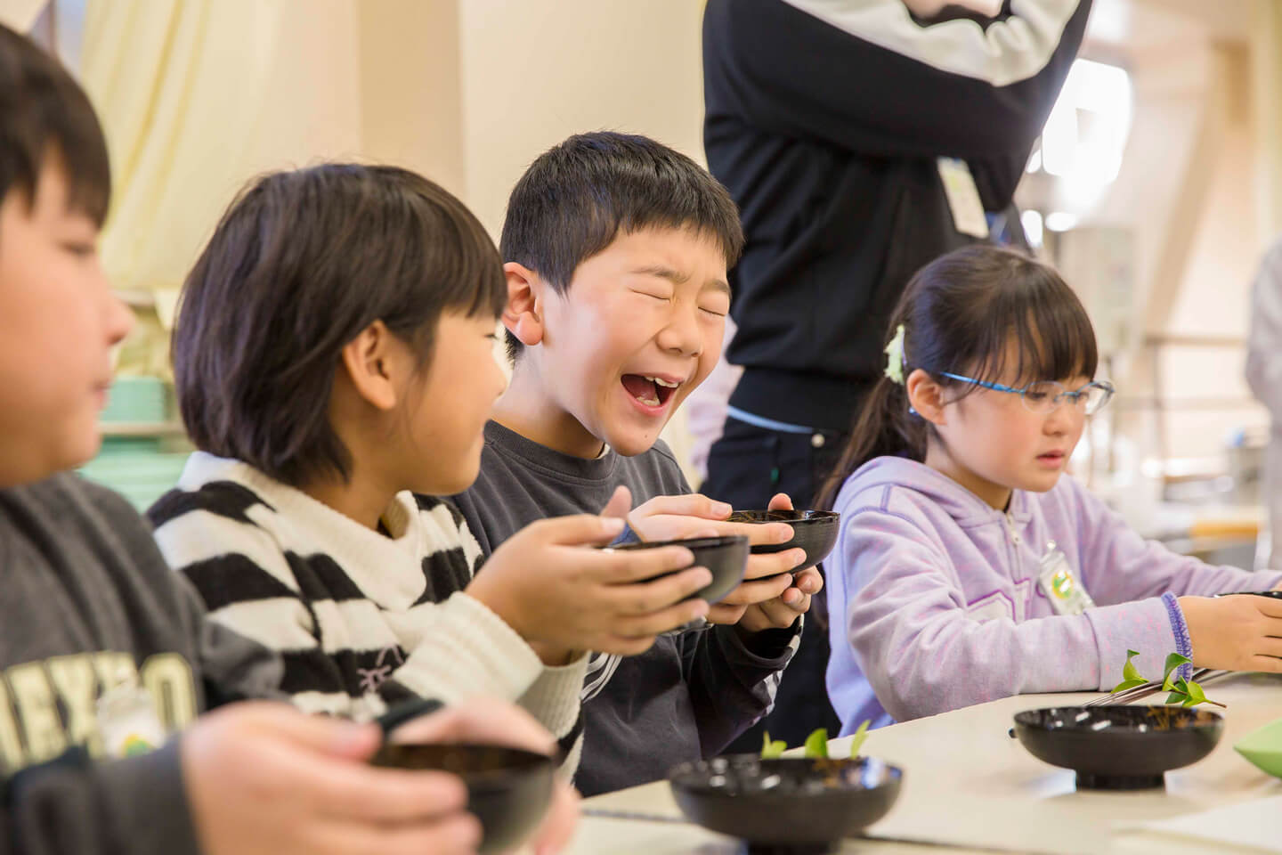 お椀の汁物を飲んだ子どもが両目を閉じて大きく口を開いた笑顔を見せている