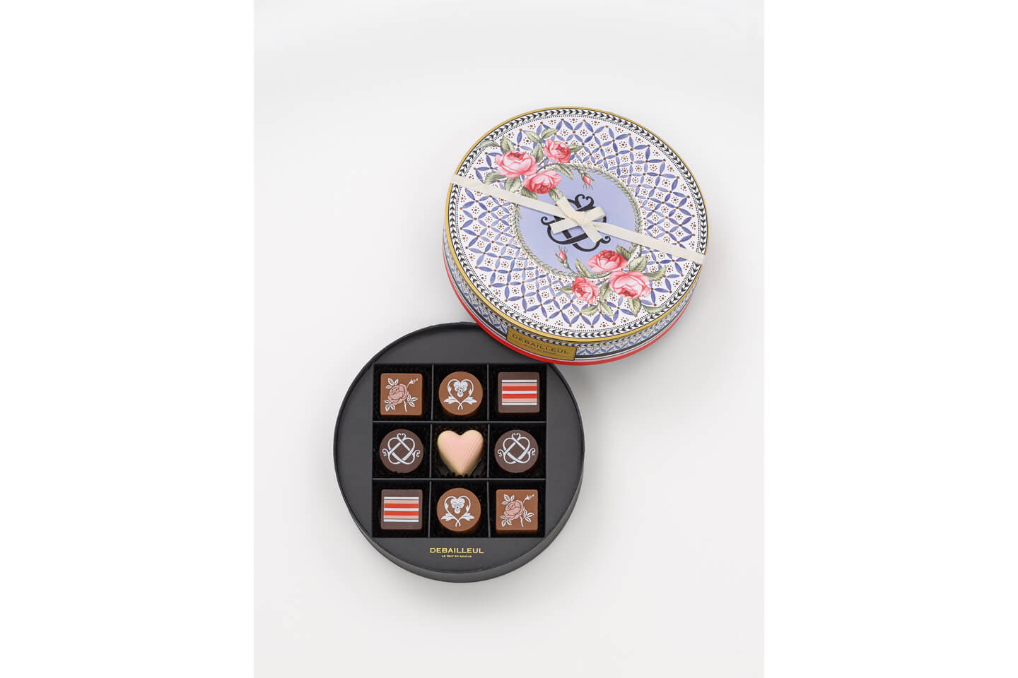 黒く丸い箱の中心に淡いピンクのはーとのチョコレート、その周囲4辺を丸いチョコレートと四角いチョコレートが囲む。チョコレートは9個入り。右斜め上に添えられた蓋には、ピンクのバラと青い幾何学模様がプリントされ、細くて白いリボンがかかっている
